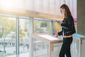 Young woman stands indoor and touches digital display while holding smartphone in her hand. Hipster...