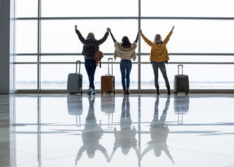 Three females standing with their backs near airport window and holding hands. Baggage is nearby