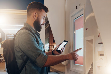 Summer night. Young hipster man with beard stands on city street and touches digital display while looks on screen of smartphone. Innovative technologies, digital display, urban navigation. Mock up.