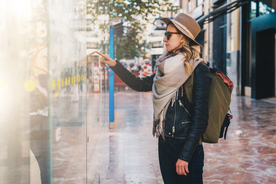 Young tourist woman in hat and with backpack stands on city street near information screen on bus stop and chooses route of trip on map. Information search, urban navigation. Travel, vacation.