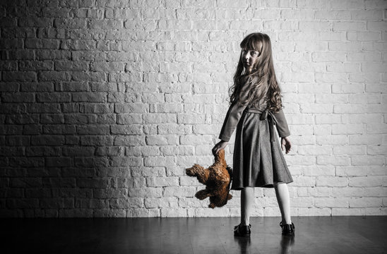 A girl with long hair stands by a white brick wall. In hand soft toy bear.
