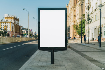 Vertical blank glowing billboard on the city street. In the background buildings and road with cars. Mock up. The poster on the street next to the roadway.