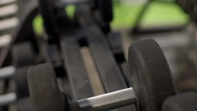 Male athlete is taking dumbbells in hands in modern gym indoors. Young bodybuilder grabs iron devices with arms in sports club. Black free weight lies on metal rack in room equipped with professional