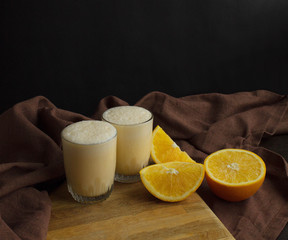 Glasses of milkshake and orange on wooden background over brown cloth