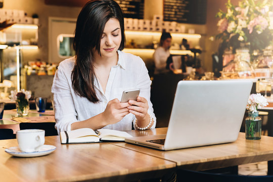 Young businesswoman sitting in cafe at table in front of laptop and using smartphone. On table notebook and cup of coffee. Online marketing, education, business planning. Distance work, e-learning.