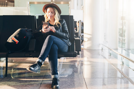 Young woman tourist in hat, with backpack sits at airport near window and uses smartphone. Hipster girl is waiting for plane landing, checks email, chatting, blogging, browsing internet.