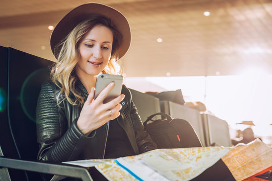 Young woman tourist in hat, with backpack sits at airport, uses smartphone, holding destination map. Hipster girl is waiting for plane landing, checks email, chatting, blogging, browsing internet.