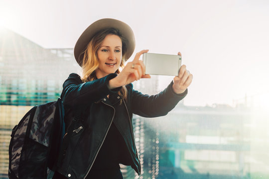 Young tourist woman in hat and with backpack stands at airport and takes photos, shoots video on smartphone's camera. Hipster girl uses digital gadget. Blurred background, bokeh effect.