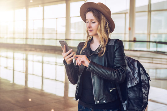 Young tourist woman in hat, with backpack stands at airport and uses smartphone.Hipster girl checks email, chatting, blogging, browsing internet.Online marketing, education. Distance work, e-learning.