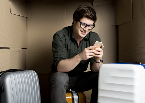 Funny news. Cheerful young man in glasses is sitting on suitcase inside confined carton box. He is holding modern mobile phone and looking at screen of device with joy