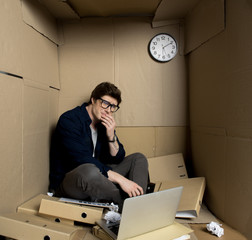 Deep depression concept. Full length of young businessman is sitting with laptop inside his small carton office and crying while touching his face. Crumpled papers and folders are scattered on floor