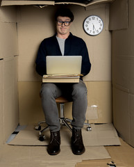 Shocked news concept. Full length portrait of surprised man in glasses is using laptop while sitting in small cardboard office. He is looking at screen of gadget with astonishment