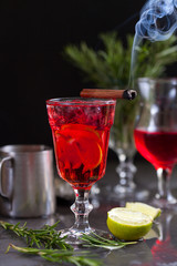 a glass with a cocktail of red color with lime, red berries, rosemary, burning cinnamon and ice cubes on a background of rosemary on a dark background