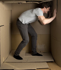 Give me more space concept. Full length of young man is standing in cardboard box and pushing wall with effort
