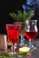 a glass with a cocktail of red color with lime, red berries, rosemary, burning cinnamon and ice cubes on a background of rosemary on a dark background