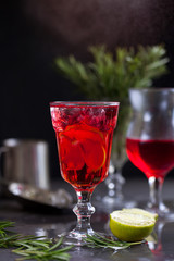 a glass with a cocktail of red color with lime, red berries, rosemary, burning cinnamon and ice cubes on a background of rosemary on a dark background