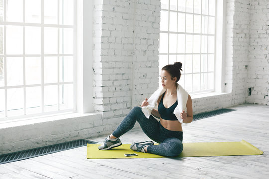 Healthy active sporty girl with hair knot exercising in light hall with large windows, resting on green mat, wiping sweat with white towel, having tired absolutely exhausted look during cardio workout
