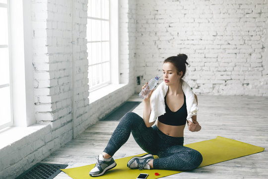 Athletic cute girl in black sports bra, leggings and running shoes relaxing after cardio training routine in gym hall, resfreshing herself with drinking water and checking email on her smart phone