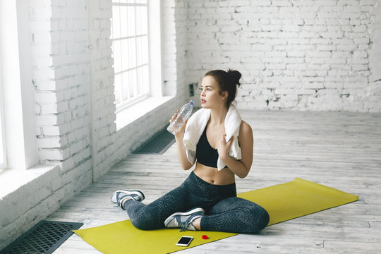 Pretty girl with perfect slim body dressed in stylish sports wear sitting on green mat in gym hall with white towel around her neck, drinking water after cardio workout, having tired expression