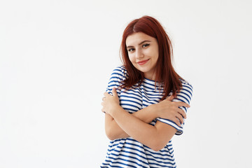 Horizontal picture of beautiful cute young 20 year old European woman in stylish sailor shirt smiling joyfully, crossing arms on her chest, embracing herself as sign of self love and self esteem