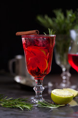 a glass with a cocktail of red color with lime, red berries, rosemary, cinnamon and ice cubes on a background of rosemary on a dark background