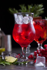 a glass with a cocktail of red color with lime, red berries, rosemary and ice cubes on a background of rosemary on a dark background