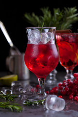 a glass with a cocktail of red color with lime, red berries, rosemary and ice cubes on a background of rosemary on a dark background