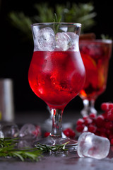 a glass with a cocktail of red color with lime, red berries, rosemary and ice cubes on a background of rosemary on a dark background