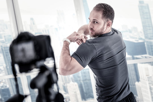 Blog for sportsmen. Bearded inspired male blogger pointing at biceps while standing in front of camera and recording video