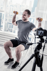 Exercises for arms. Energetic enthusiastic male blogger lifting dumbbells while sitting on chair and recording video