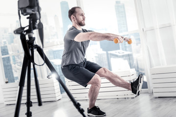 Obraz na płótnie Canvas Healthy lifestyle. Appealing pleasant male blogger using dumbbells while rising leg and recording video