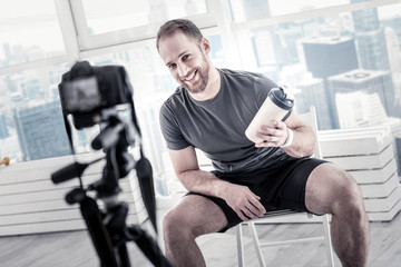 After training program. Optimistic vigorous male blogger showing bottle while holding it and smiling