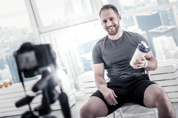 After exercise session. Cheerful gay male blogger carrying bottle while grinning and sitting on chair