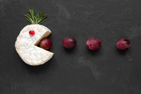Camembert or brie cheese cut out like pacman eating grapes on black slate background
