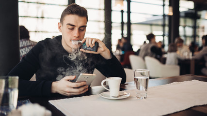 Young man using electronic cigarette to smoke in closed public space.Satisfied e- cigarette user in cafe.Smoking ban alternative,nicotine addiction,quitting tobacco concept.Inhaling flavor