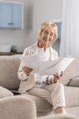 Accurate report. Happy jolly mature businesswoman posing on couch while scrutinizing papers and smiling