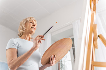 Creating process. Low angle of Appealing pleasant mature woman carrying palette and tassel while staying and searching inspiration