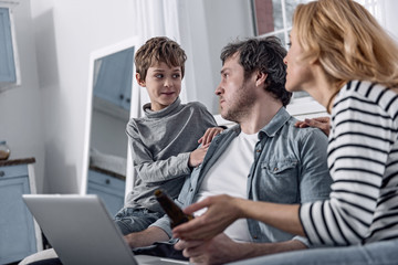 Fototapeta na wymiar Cheer up. Kind attentive cheerful boy looking at his exhausted depressed father holding a modern laptop while sitting on a sofa with him and mother