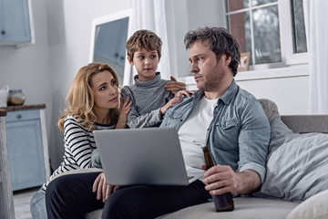 Fototapeta na wymiar Kind relatives. Exhausted depressed alcoholic man sitting at home with a modern laptop on his knees and a bottle in his hand while a kind loving wife hugging their son and looking at him