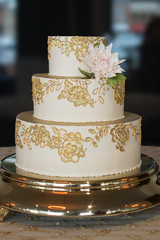 3 Tier Wedding Cake With Gold Embroidery And A Single Edible White Dahlia Flower On Top Of A Gold Cake Stand