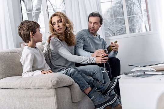 Finding solution. Calm upset young woman fighting her alcohol addiction and looking at the little kind boy near while her calm serious husband taking a glass of wine from her hand
