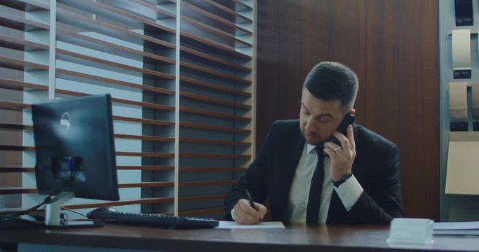 Busy adult agent in suit talking on phone while checking computer and writing in paper in office.