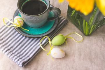 Fototapeta na wymiar Top view of a gray and blue cup of black coffee, yellow daffodils flowers in a vase, Easter eggs decoration on natural cotton cloth background. Easter morning or breakfast background, vintage toned