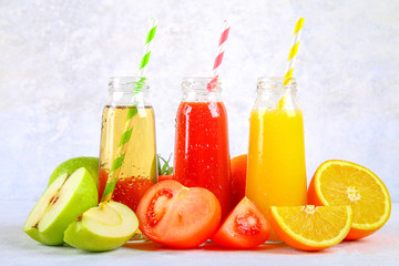 Bottles with fresh orange, apple, tomato juice and colored tubules on a gray concrete table.