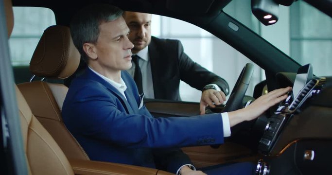 Side view of seller standing at window and showing options to adult businessman using car computer.