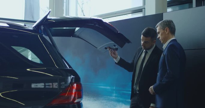 Side view of manager in suit showing car trunk size to adult businessman in showroom.