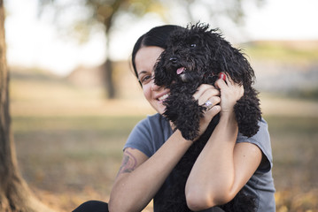 Young woman cuddling with her cute little miniature black poodle, sitting together in a park, enjoying the sunny and warm weather