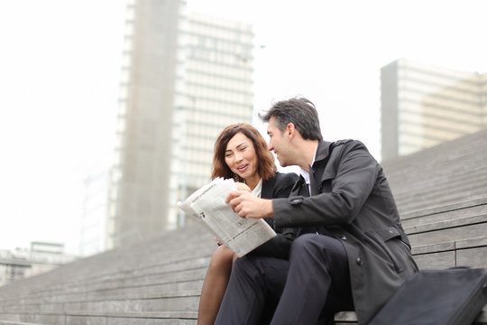 Engineers Male And Female Find Article About Company In Newspaper, Colleagues Sitting On Stairs Reading. Americans Wearing Business Clothes Smiling Communicating. Concept Of Fashionable Cl