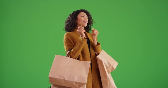 Fashionable woman dancing while holding shopping bags on greenscreen background. Stylish black millennial shopper dances in yellow coat on green screen for keying or compositing. 4k