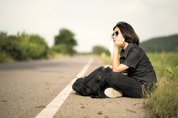 Woman sit with backpack hitchhiking along a road in countryside